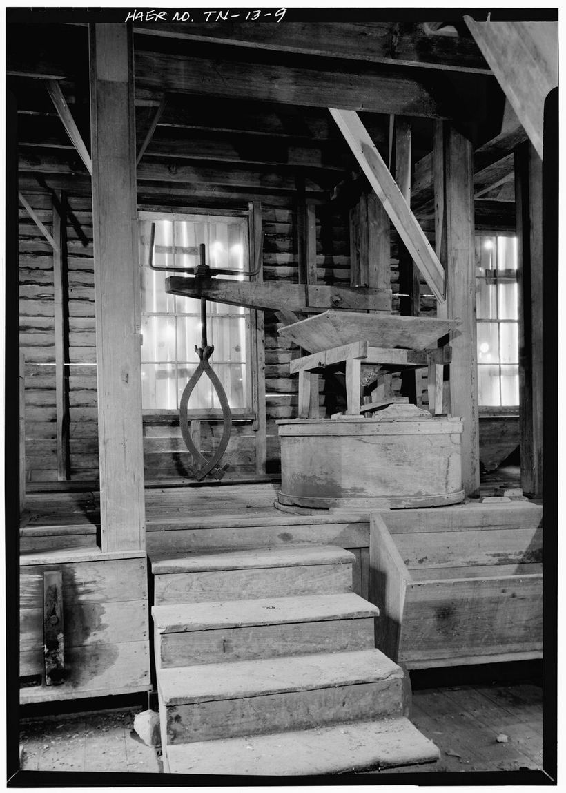 Inside a stone mill: a wooden hopper and shoe above a round stone case, with a tentering screw
          on the left and log walls behind.